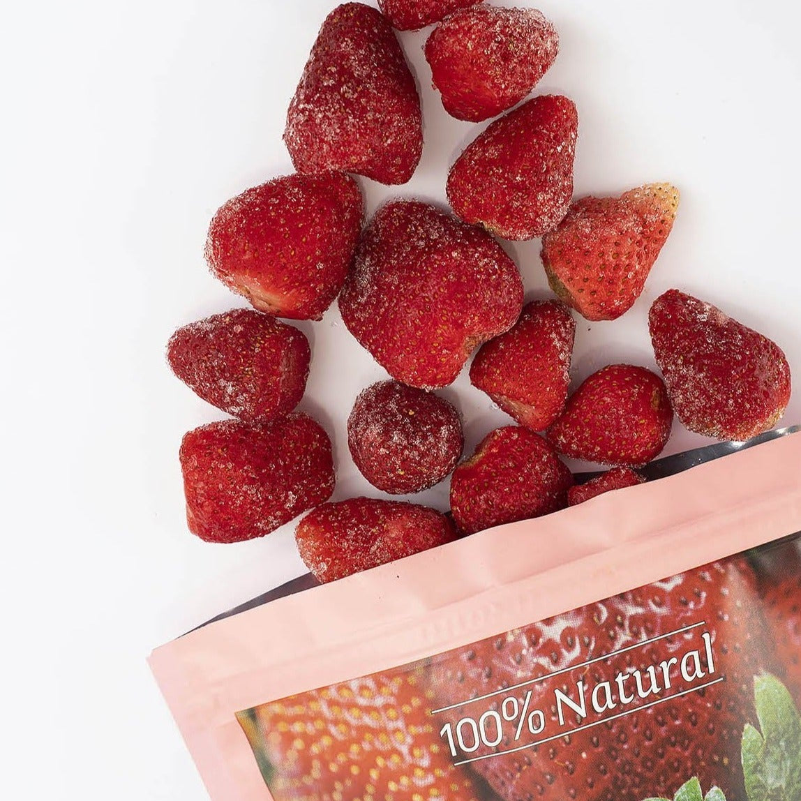 Frozen red strawberries piled on top of a pink stand-up pouch labeled '100% Natural' on a white background.
